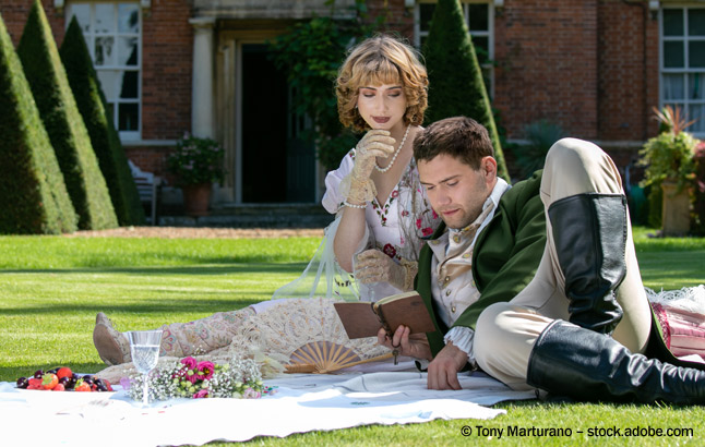 Young lovers dressed in vintage clothing sitting on picnic blanket. Gentleman reading to his lover from a book of poems 